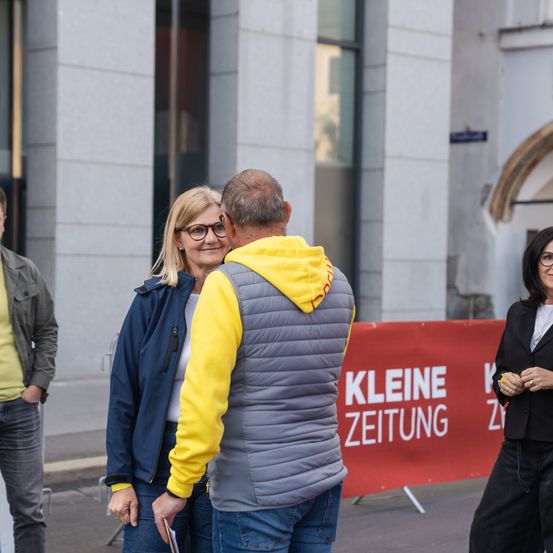 Ein Mann und eine Frau stehen draußen, lächeln und unterhalten sich. Eine Frau rechts beobachtet sie, und ein Mann steht in der Nähe eines Gebäudes.