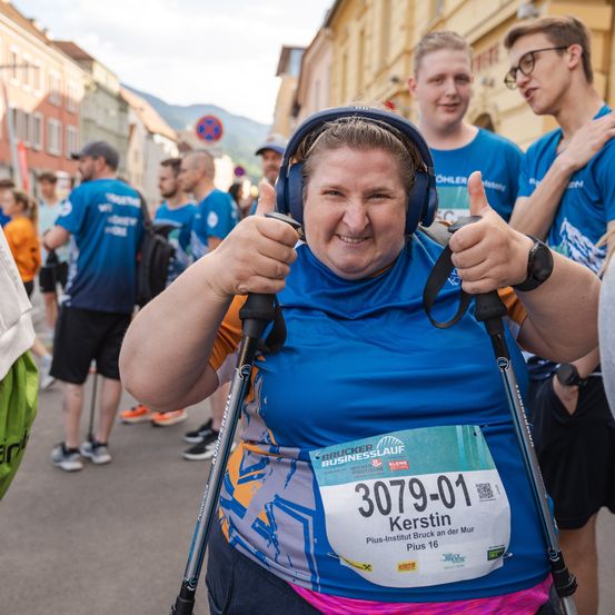 Eine lächelnde Frau mit einem Läufer-Startnummernschild auf ihrem Shirt gibt ein Daumen hoch, während sie Skistöcke hält. Hinter ihr stehen mehrere Leute mit blauen Shirts auf einer Stadtstraße.