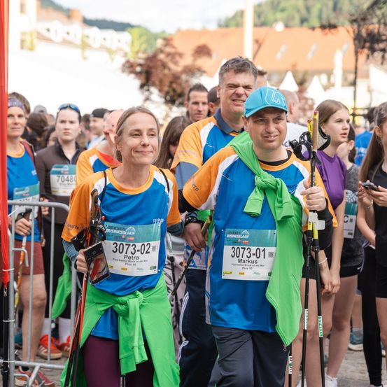 Zwei Läufer, Petra und Markus, stehen lächelnd in einem Marathon. Petra trägt einen grünen Umhang und eine Nummer 3073-02, Markus trägt einen blauen Hut und Umhang 3073-01. Sie halten Skistöcke. Hinter ihnen versammelt sich eine Menge.