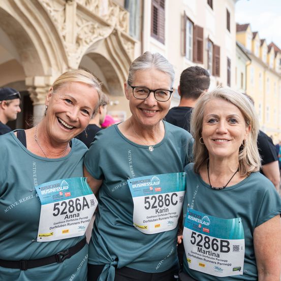Drei Frauen mit Laufnummern stehen zusammen und lächeln für die Kamera. Dahinter befindet sich ein historisches Gebäude mit Bögen.