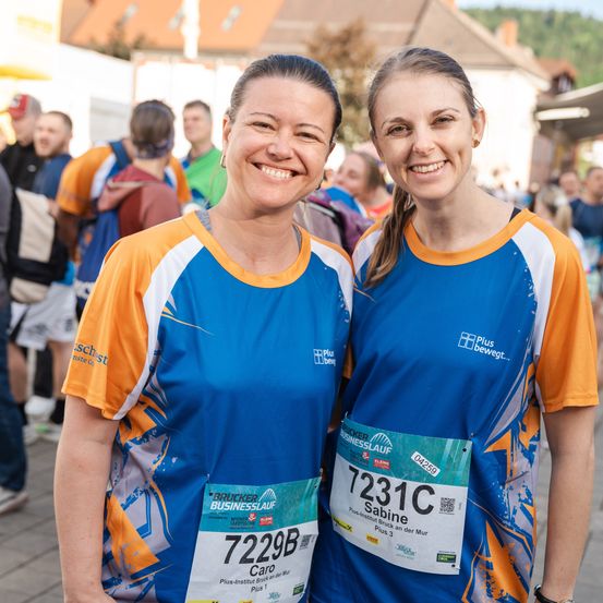 Zwei Frauen in passenden blauen und orangefarbenen Shirts lächeln in die Kamera und stehen zusammen in einem überfüllten Außenbereich, wahrscheinlich bei einer Marathonveranstaltung.