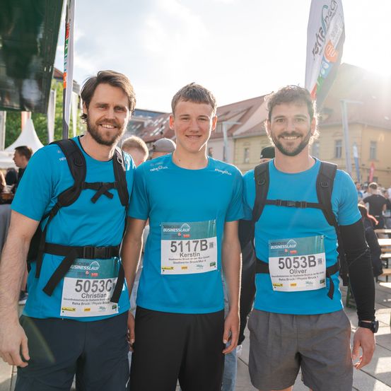 Drei Männer in blauen Shirts und Rucksäcken posieren für ein Foto. Der Mann in der Mitte hat die Nummer 5117B auf seinem Shirt, während der Mann auf der rechten Seite 5053B hat. Sie befinden sich bei einem Marathon-Event.