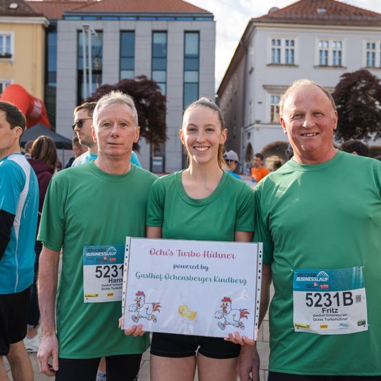 Drei Personen stehen vor einem Gebäude und lächeln für die Kamera. Der Mann auf der linken Seite trägt ein blaues Shirt, die anderen grüne T-Shirts. Die Frau hält eine Mappe mit dem Titel 'Ochis Turbo Hühner.' Sie haben Startnummern: 5231, 5231B und 5231 Fritz.