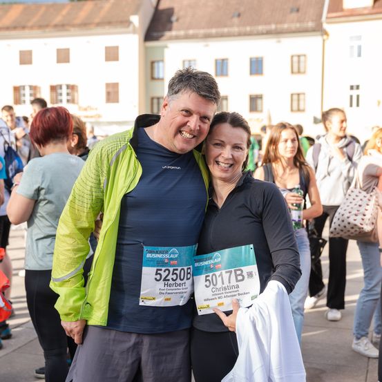 Ein Mann und eine Frau stehen lächelnd mit Laufnummern vor einem Gebäude. Sie sind von anderen Leuten umgeben.