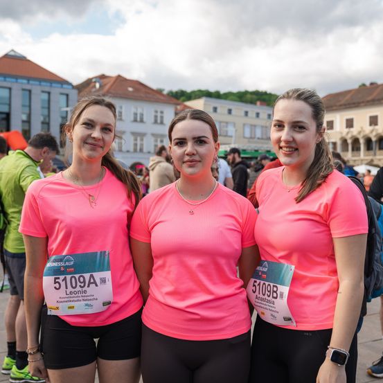 Drei Frauen in rosa Shirts mit den Nummern 5109A, 5109B und 5109C stehen nebeneinander vor einer verschwommenen Menge.