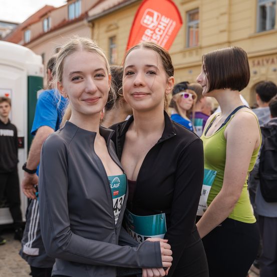 Zwei lächelnde Frauen in Laufbekleidung stehen zusammen in einer Menschenmenge, eine trägt die Nummer 43. Dahinter sind ein Banner und Gebäude zu sehen, was auf einen Laufwettbewerb hinweist.