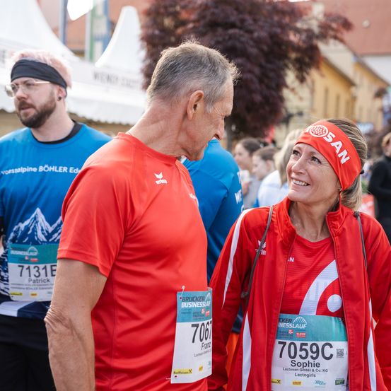 Marathonteilnehmer in roten Shirts stehen zusammen. Ein Mann vorne trägt die Nummer 7067, und eine Frau neben ihm hat eine Stirnband mit "OSSAN" und die Nummer 7059C.