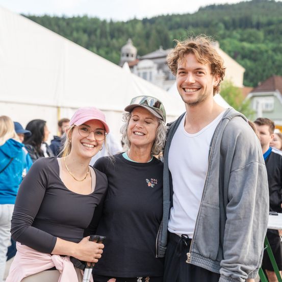 Drei Personen posieren für ein Foto bei einer Outdoor-Veranstaltung. Die Frau links trägt eine rosa Kappe und eine Brille. Die mittlere Frau hat ein schwarzes Shirt und eine Kappe. Der Mann rechts trägt eine graue Hoodie. Sie alle lächeln für die Kamera.