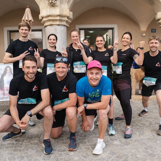 Eine Gruppe von Läufern in passenden schwarzen und blauen T-Shirts posiert für ein Foto vor einem Gebäude. Einige knien, andere stehen, alle lächeln mit Friedenszeichen.