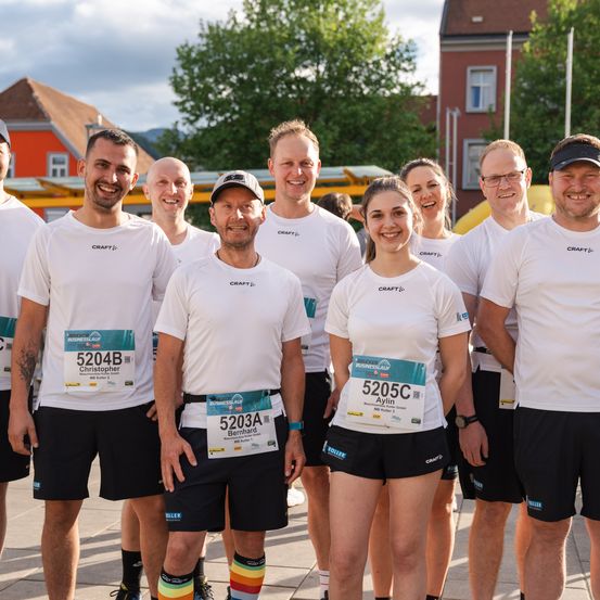 Eine Gruppe von Läufern in weißen Craft-T-Shirts und schwarzen Shorts posiert für ein Foto, mit auf ihren Shirts angebrachten Startnummern. Sie stehen vor Gebäuden und Bäumen.
