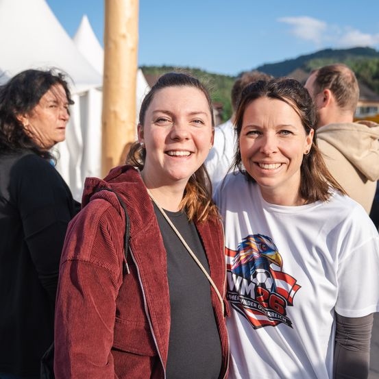 Zwei lächelnde Frauen stehen nebeneinander, mit anderen im Hintergrund. Die Frau links trägt eine rote Jacke, und die auf der rechten Seite trägt ein weißes T-Shirt mit einem Fußballteam-Logo.