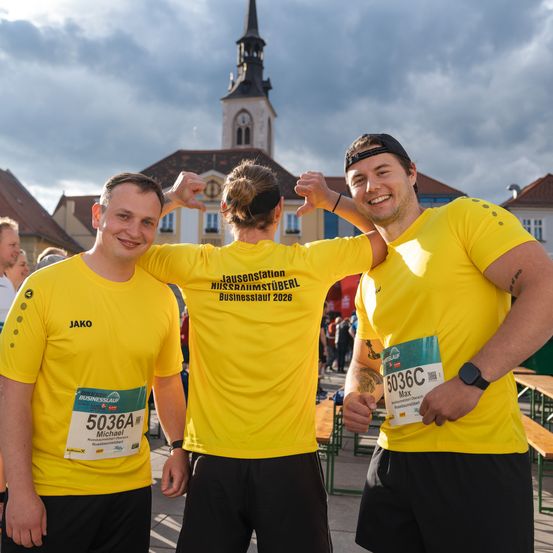 Drei Menschen in gelben T-Shirts mit Text und Zahlen stehen vor einem Gebäude und lächeln. Ein Kirchturm ist im Hintergrund.