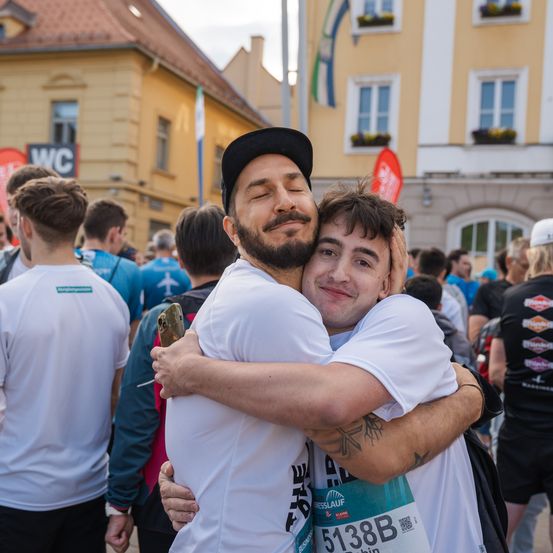 Läufer in weißen T-Shirts umarmen sich inmitten einer Menschenmenge bei einem Marathon. Im Hintergrund sind Gebäude mit Fahnen und Bannern zu sehen.