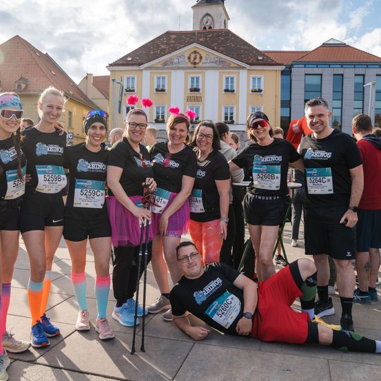 Läufer posieren für ein Foto bei einem Marathon-Event, tragen nummerierte Startnummern und verschiedenfarbige Socken. Ein Mann liegt vor ihnen auf dem Boden.