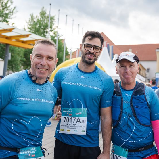 Drei Männer in blauen Shirts posieren für ein Foto, wahrscheinlich nach einem Marathon. Der Mann in der Mitte trägt eine Startnummer 7017A. Sie stehen auf einem Betonuntergrund mit einem Gebäude und Bäumen im Hintergrund.