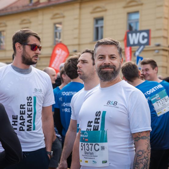 Eine Gruppe von Läufern mit nummerierten Startnummern nimmt an einem Marathon teil. Ein Läufer trägt ein weißes Shirt mit The Paper Makers darauf.