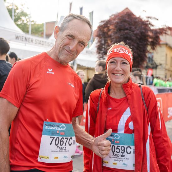 Ein lächelnder Mann und eine Frau in passenden roten Shirts posieren für ein Foto bei einem Marathon-Event. Der Mann trägt Franz' Startnummer 70670 und die Frau trägt Sophies Nummer 059C.
