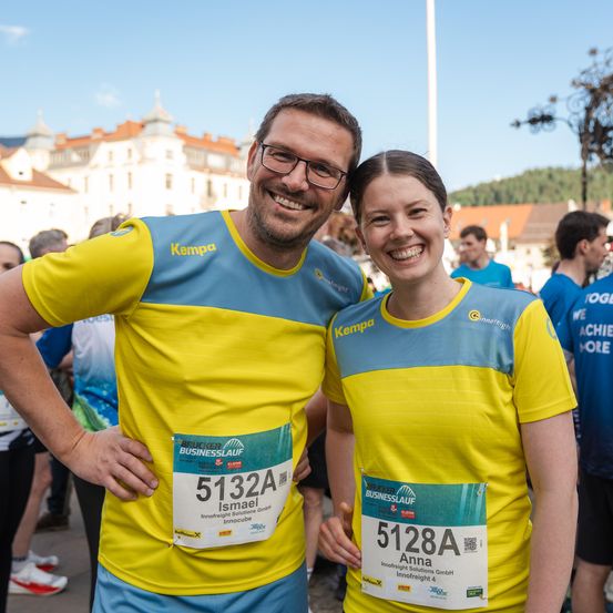 Ein Mann und eine Frau stehen zusammen und tragen passende gelbe und blaue T-Shirts mit Nummern. Sie lächeln für die Kamera. Dahinter sind verschwommene Bilder von Menschen und Gebäuden.
