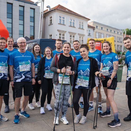 Eine Gruppe von Menschen in blauen T-Shirts posiert für ein Foto, lächelt und hält Wanderstöcke. Sie sind wahrscheinlich Teilnehmer eines Marathons. Im Hintergrund befinden sich Gebäude mit Glasfenstern und ein gelber Banner.