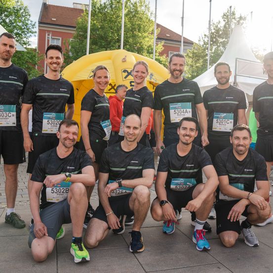 Eine Gruppe von Menschen in einheitlichen Outfits posiert für ein Foto im Freien. Sie alle lächeln und haben Nummern auf ihren Trikots. Hinter ihnen befinden sich Zelte und Bäume.
