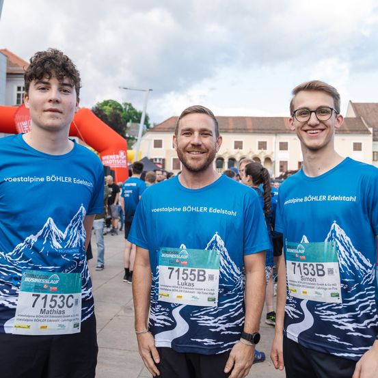 Drei Männer in blauen T-Shirts lächeln in die Kamera. Sie stehen vor einem Gebäude mit einem orangefarbenen Bogen. Der Mann in der Mitte hat eine Nummer 7155B auf seinem Shirt.