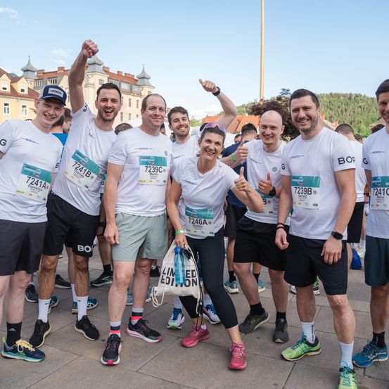 Eine Gruppe von Marathonläufern mit Nummernschildern steht und posiert für ein Foto. Sie tragen alle weiße Hemden und Shorts. Einige von ihnen lächeln und einer von ihnen hält eine Tasche. Der Mann in der Mitte trägt einen Hut und hat die Hände hoch. Hinter ihnen ist ein verschwommener Hintergrund mit einem Gebäude und Bäumen.
