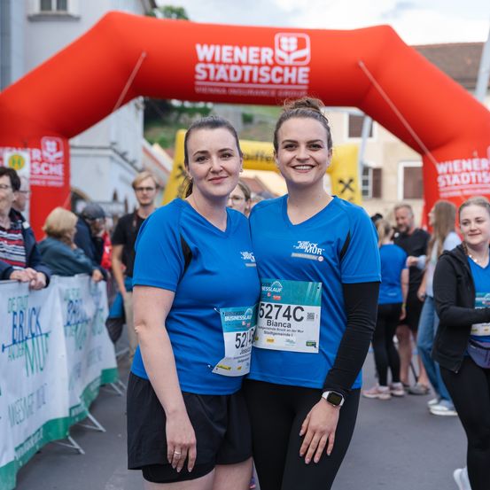 Zwei Frauen in blauen Shirts stehen vor einem roten Torbogen mit einem Marathonlogo und lächeln für die Kamera. Hinter ihnen schaut eine Menschenmenge zu, einige tragen Brillen. Der Torbogen steht 'Wiener Stadtische'.