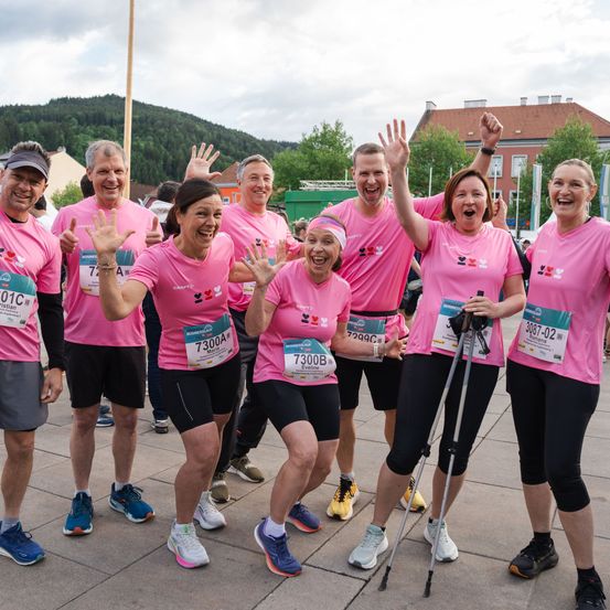 Eine Gruppe von Läufern, alle in pinkfarbenen T-Shirts, stehen zusammen und lächeln für ein Foto. Die Frauen wedeln mit den Händen, und der Mann rechts hat eine Krücke. Sie sind in einem Außenbereich mit Gebäuden und Bäumen in der Ferne.