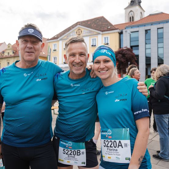 Drei Personen in türkisfarbenen Sporttrikots stehen lächelnd vor einem Gebäude. Sie haben Nummern auf ihren Trikots, was darauf hindeutet, dass sie an einem Marathon teilnehmen.