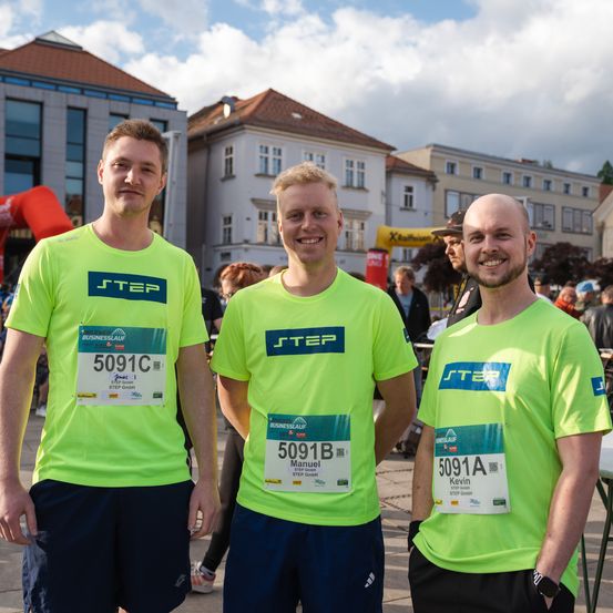 Drei Männer in neongrünen T-Shirts mit Nummern darauf lächeln und posieren für ein Foto vor einem Gebäude.