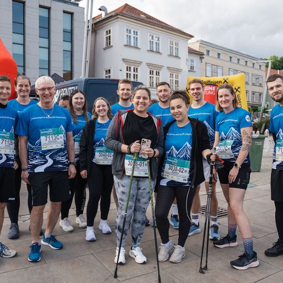 Eine Gruppe von Menschen, alle mit passenden blauen Shirts und Turnschuhen bekleidet, posiert mit ihren Wanderstöcken in der Hand vor einem Gebäude für ein Foto.