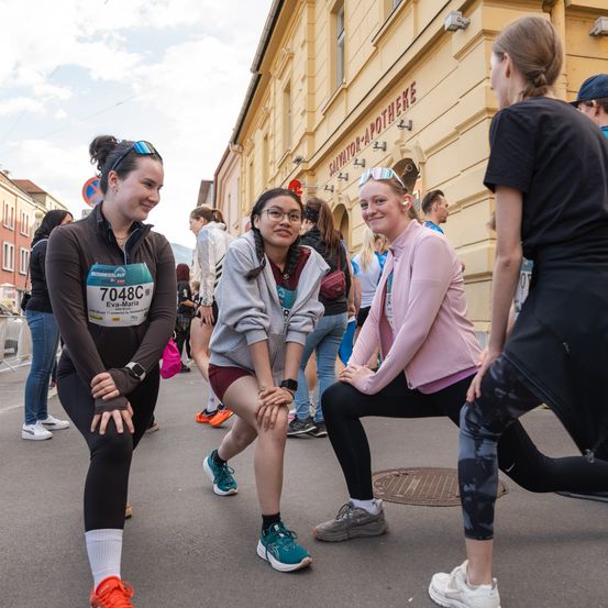 Drei Frauen hockten auf einer Straße, wahrscheinlich als Teilnehmer eines Marathons. Sie sind von anderen Teilnehmern und Zuschauern umgeben. Ein Gebäude mit einem roten Schild ist im Hintergrund.