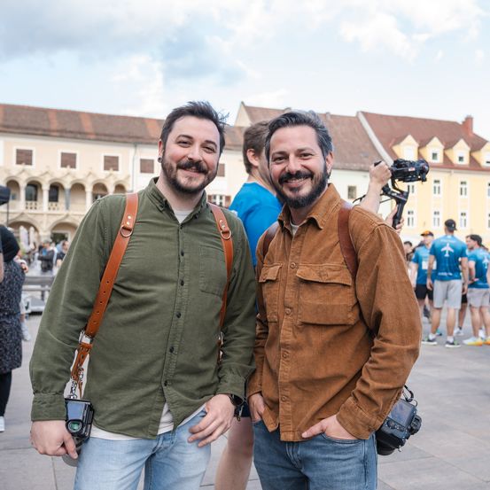 Zwei Männer mit Kameras stehen vor einem historischen Gebäude. Sie tragen lässige Kleidung und lächeln für die Kamera. Hinter ihnen gehen Leute umher und ein Mann mit einer Kamera auf einem Stativ hält die Szene fest.