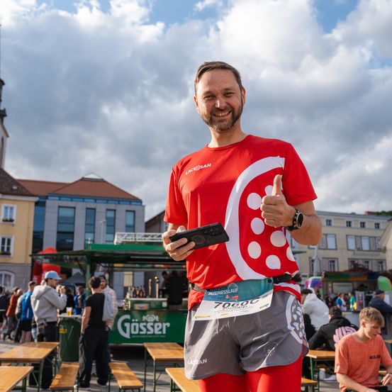 Ein lächelnder Mann in einem roten Shirt und Laufausrüstung zeigt den Daumen nach oben vor einer überfüllten Straße mit einem Imbisswagen und Gebäuden im Hintergrund.