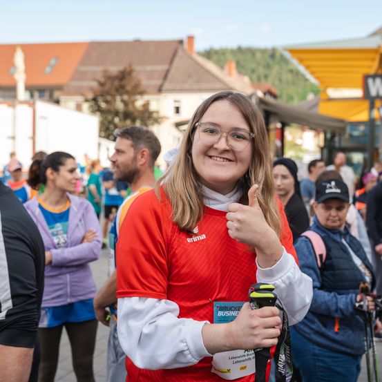 Eine Frau in einem roten Shirt gibt bei einem Rennen ein Daumen hoch, mit vielen Menschen im Hintergrund.