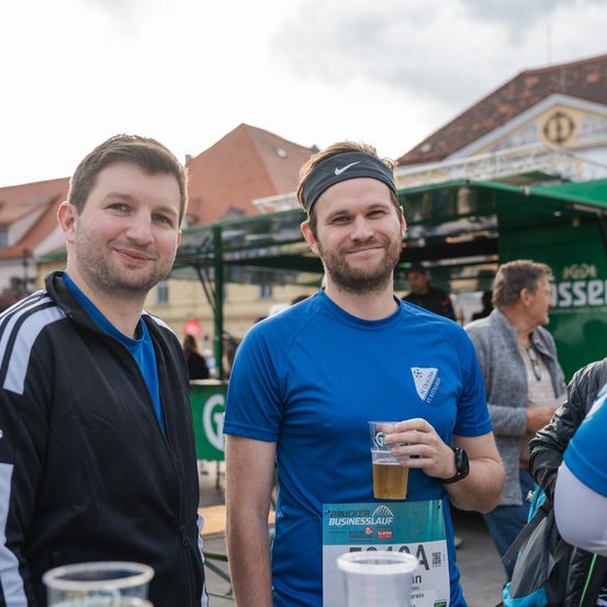 Zwei Männer stehen lächelnd für ein Foto, einer trägt ein blaues T-Shirt mit einem Becher in der Hand und eine schwarze Kopfband. Der andere in einem schwarzen und weißen Jacke hält einen Becher. Dahinter ist eine grüne Struktur mit Text.