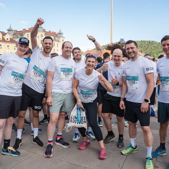Eine Gruppe von Läufern in gleichen weißen Shirts posiert vor einem Marathon. Die Frau in der Mitte hält eine Wasserflasche. Hinter ihnen steht ein Gebäude mit Türmen.