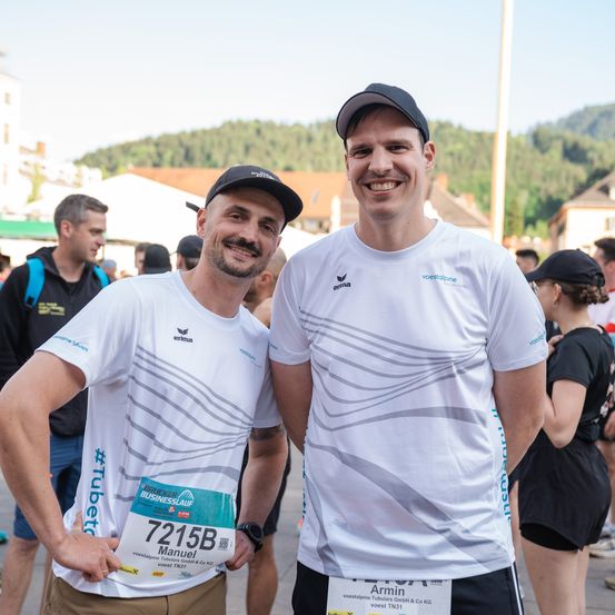 Zwei Männer stehen bei einem Marathon zusammen und lächeln für die Kamera. Sie tragen passende weiße T-Shirts mit Logos und Nummern, einer mit der Nummer 7215B. Hinter ihnen sind verschwommene Figuren von anderen Teilnehmern und Zuschauern zu sehen.