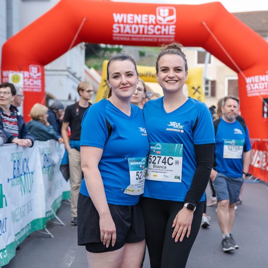 Zwei Frauen stehen vor einem roten Bogen bei einem Marathon. Sie tragen blaue Shirts mit Nummern und lächeln. Zuschauer und ein Banner mit Text sind hinter ihnen.