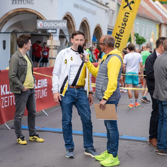 Ein Mann in einer weißen Jacke wird von einem anderen Mann in einer gelben und grauen Jacke bei einer Outdoor-Veranstaltung interviewt. Sie stehen vor einem gelben Banner, mit einer Menge und Gebäuden im Hintergrund.