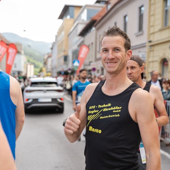 Ein Mann in einem schwarzen Tanktop gibt ein Daumen hoch auf einer Menschen- und Autoleeren Straße. Gebäude und Berge sind im Hintergrund sichtbar.