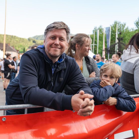 Ein Mann und ein Junge lächeln bei einer Laufveranstaltung. Sie sind von Zuschauern umgeben, einer davon ist eine Frau mit Pferdeschwanz. Die Veranstaltung findet im Freien statt, mit Bergen im Hintergrund.