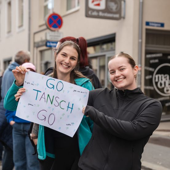 Zwei junge Frauen halten ein Schild mit der Aufschrift Go Tansch Go auf einer überfüllten Straße und lächeln. Sie sind von Menschen und Gebäuden umgeben.