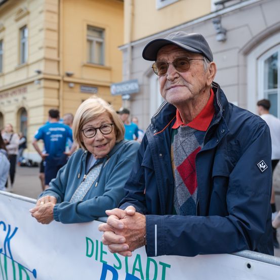 Ein älteres Paar steht neben einem Banner und lächelt. Das Banner steht für 'Die Stadt'. Sie befinden sich bei einer Veranstaltung im Freien.