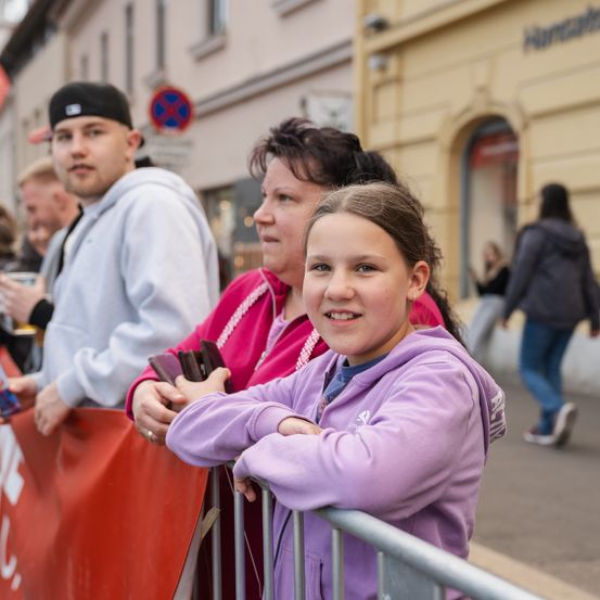 Eine Gruppe von Menschen steht hinter einem Zaun bei einer Veranstaltung im Freien. Ein Mädchen in einer lila Jacke lächelt. Ein Mann mit schwarzem Hut und eine Frau in einer rosa Jacke stehen hinter ihr.