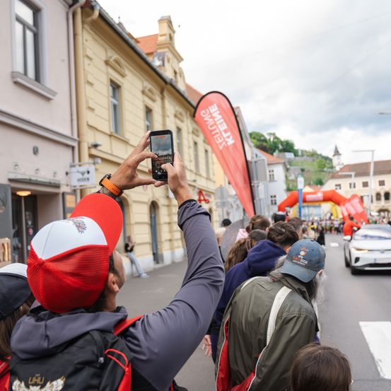 Ein Mann mit rotem Hut fotografiert ein vorbeifahrendes Auto bei einer Straßenveranstaltung. Im Hintergrund sind ein Banner und Gebäude zu sehen.