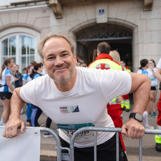 Ein Mann in einem weißen T-Shirt mit der Nummer 72 steht lächelnd hinter einem Barriere und trägt eine Smartwatch und einen Ring. Dahinter befinden sich Personen in Sportbekleidung, einige mit roten und gelben Westen. Im Hintergrund steht ein Gebäude.