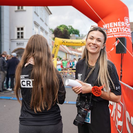 Zwei Frauen in schwarzen T-Shirts stehen neben einem Bogen. Eine Frau hält eine Kamera und ein gerolltes Papier. Dahinter versammelt sich eine Menge. Der Bogen trägt die Aufschrift 'Wiener Hadtische'.