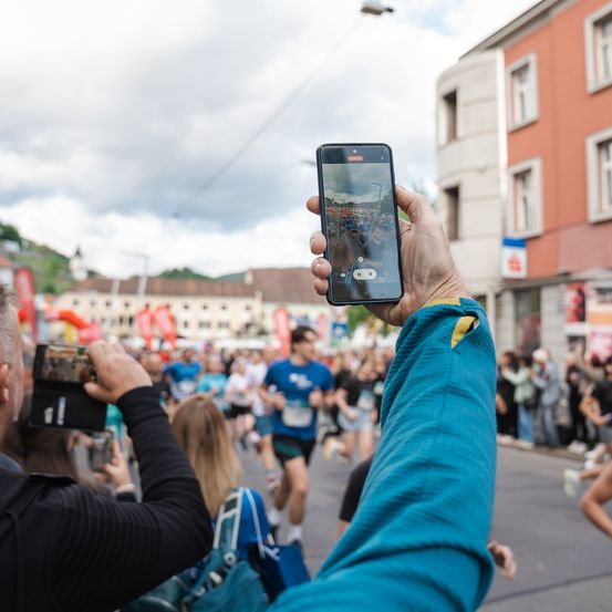 Ein Mann fotografiert einen Marathon mit seinem Smartphone, mit Läufern und Zuschauern im Hintergrund. Die Menge steht entlang der Straße und beobachtet das Ereignis.