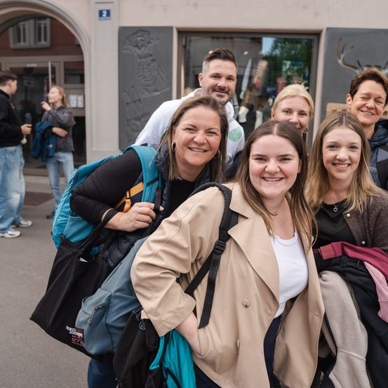 Eine Gruppe lächelnder Menschen steht vor einem Gebäude. Sie tragen Jacken und haben Taschen bei sich, wobei eine Frau eine Halskette trägt. Hinter ihnen halten zwei Personen Jacken, und eine Person trägt einen Rucksack. Im Hintergrund sind Gebäude mit Glasfenstern zu sehen.